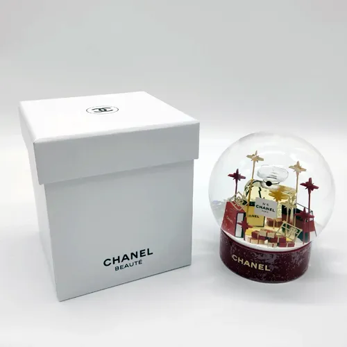 Chanel. Boule à neige figurant un flacon de parfum Chanel N°5 entouré de paquets cadeaux et d'étoiles fixées sur tiges dorées et rouge, H.11,5 cm. Montée sur socle rouge titré Chanel.